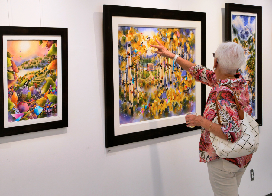 A gallery launch attendee pointing to one of Alex Fong's paintings that shows a vibrant field with flowers and a lake in the background.