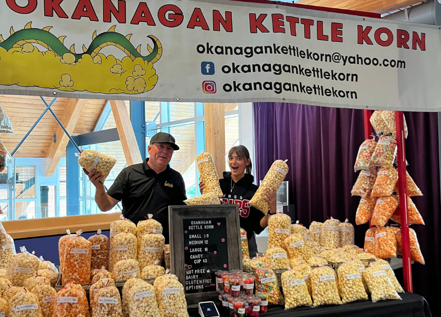 Okanagan Kettle Corn staff pose behind their stands, popcorn bags in hand smiling.